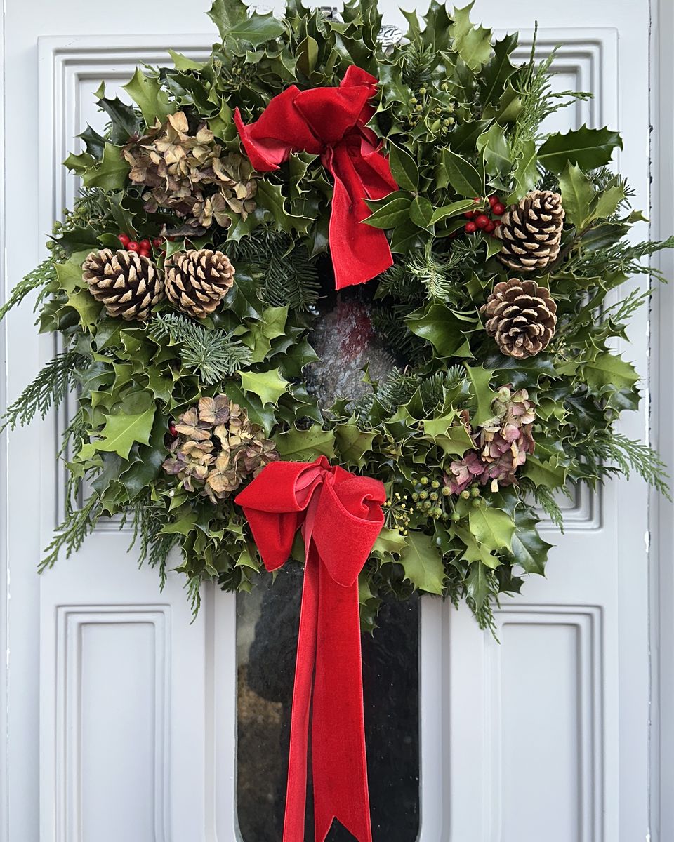 A fresh holly wreath with pine cones and a red velvet ribbon on a white front door